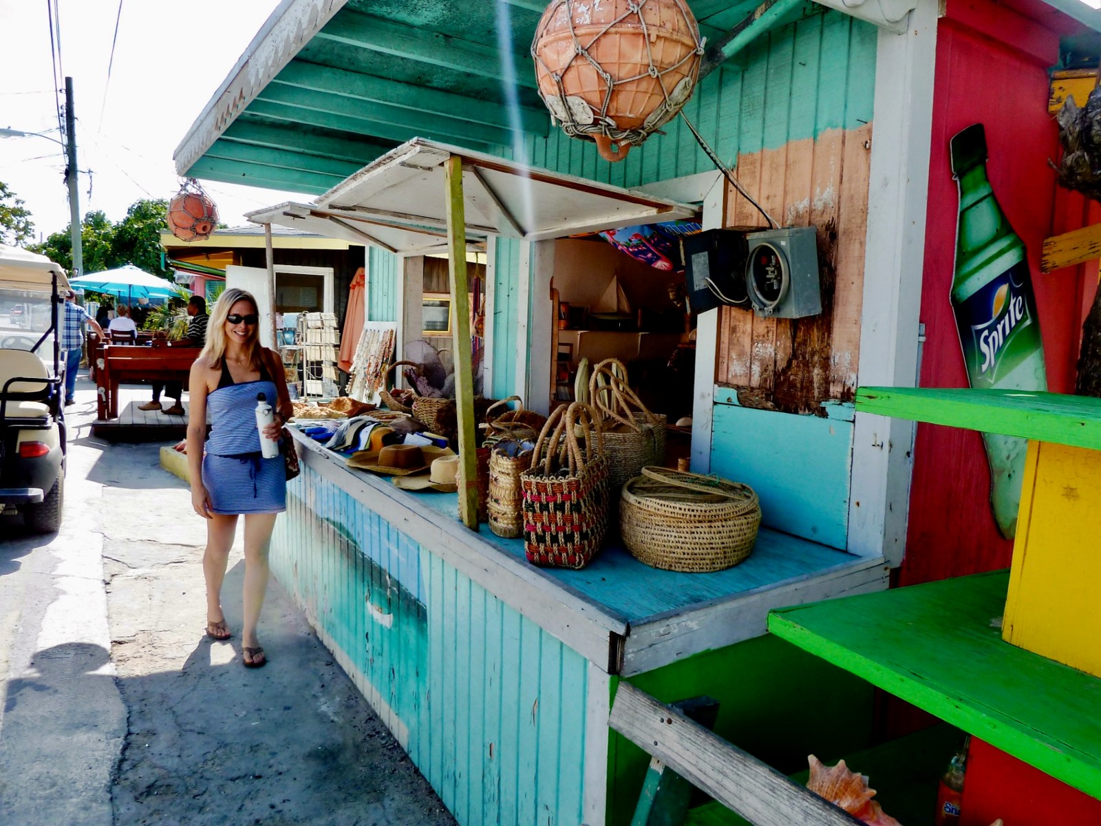 Julie walking past a colorful Bahamian straw market stall with woven baskets and sun hats