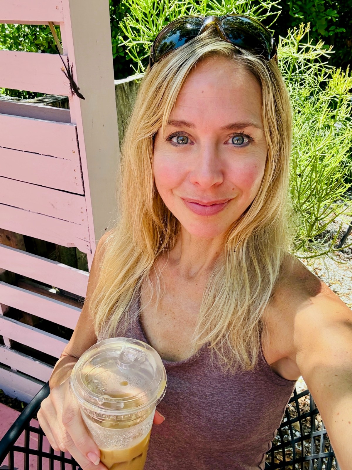 Julie smiling outdoors with an iced coffee beside a pink wooden gate and tropical greenery
